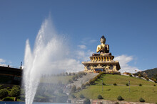 Buddha Statue With Blue Sky And Clouds In The Background And A Water Fountain Throwing Water Upwards  In The Foreground 