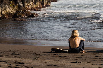 Young surfer man sitting on the beach with board after surf session at summer sunset - Focus on back