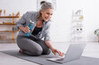 © LIGHTFIELD STUDIOS - smiling middle aged woman sitting on fitness mat, holding sports bottle and using laptop