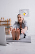 © LIGHTFIELD STUDIOS - smiling mature woman with grey hair stretching on yoga mat near laptop on blurred foreground