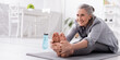 © LIGHTFIELD STUDIOS - happy mature woman with grey hair stretching on yoga mat near sports bottle, banner