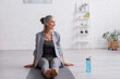 © LIGHTFIELD STUDIOS - happy mature woman with grey hair sitting on yoga mat near sports bottle