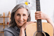 © LIGHTFIELD STUDIOS - happy mature woman with grey hair holding acoustic guitar in living room