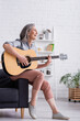 © LIGHTFIELD STUDIOS - cheerful mature woman with grey hair playing acoustic guitar while sitting in living room