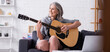 © LIGHTFIELD STUDIOS - pleased mature woman with grey hair learning to play acoustic guitar near laptop, banner