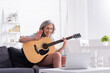 © LIGHTFIELD STUDIOS - middle aged woman with grey hair holding acoustic guitar while waving hand during video call on laptop
