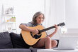 © LIGHTFIELD STUDIOS - happy middle aged woman learning to play acoustic guitar near laptop on coffee table
