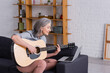 © LIGHTFIELD STUDIOS - happy middle aged woman with grey hair learning to play acoustic guitar near laptop on sofa