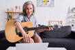 © LIGHTFIELD STUDIOS - middle aged woman with grey hair learning to play acoustic guitar while reaching laptop