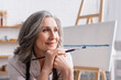 © LIGHTFIELD STUDIOS - happy middle aged painter with grey hair holding paintbrush while looking away