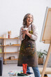 © LIGHTFIELD STUDIOS - excited middle aged artist holding cup of coffee and paintbrush near canvas
