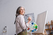 © LIGHTFIELD STUDIOS - cheerful middle aged woman holding paintbrush and palette with colorful paints near blank canvas