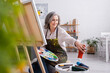 © LIGHTFIELD STUDIOS - mature woman holding paintbrush and palette while reaching red paint in tube and sitting near canvas