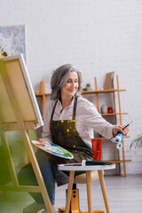 pleased mature artist holding paintbrush and palette while reaching blue paint in tube and sitting near canvas