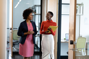 Happy african american female business colleagues walking through corridor holding documents talking