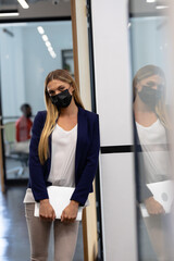 caucasian businesswoman wearing mask standing in corridor holding laptop looking to camera
