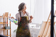 © LIGHTFIELD STUDIOS - cheerful mature woman in apron holding paintbrush and palette near easel at home