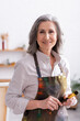 © LIGHTFIELD STUDIOS - cheerful middle aged artist in apron with spills holding paintbrush
