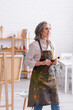 © LIGHTFIELD STUDIOS - pleased middle aged artist in apron with spills holding paintbrush at home