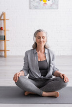Pleased Middle Aged Woman In Wireless Headphones Meditating While Sitting In Lotus Pose On Yoga Mat At Home