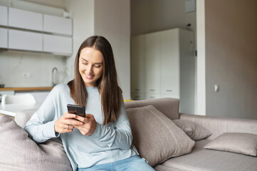 Wall Mural - Serene young woman holds smartphone sitting on the couch at home, chatting online with friends, spend leisure time in social media, texting, scrolling news feed, shopping online