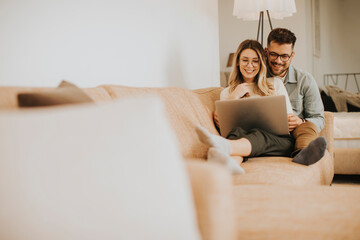 young couple using laptop together while sitting on sofa at home