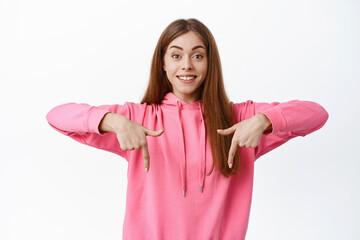 Wall Mural - Excited young woman shows advertisement on bottom, pointing fingers down at copy space logo, smiling happy, showing promo deal, white background