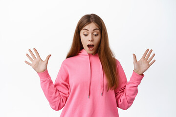 Wall Mural - Image of young woman gasping surprised, spread hands sideways and look down and screaming, standing against white background