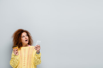 Wall Mural - surprised woman looking away while holding smartphone and credit card isolated on grey