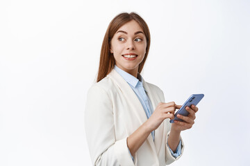 Wall Mural - Smiling corporate girl in suit using mobile phone, turn head behind and look at promotional text, reading copy space on white background, holding smartphone