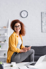 Wall Mural - happy freelance looking at camera while sitting on desk with smartphone near laptop