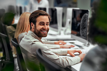 Handsome business man works at the computer with colleagues in the office and smiles looking at the camera. Data analysis, statistics, finance. Training, business team, programming, development.