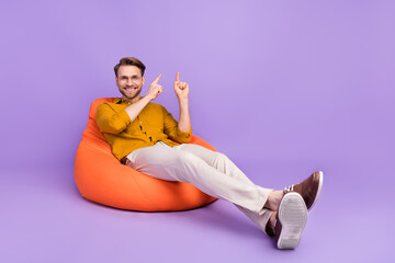 Sticker - Portrait of attractive cheerful guy sitting on chair showing copy empty space isolated over violet purple color background