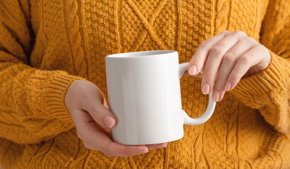 Female hands hold mock up white empty mug, cup for your design and logo close-up. Woman in yellow knitted sweater autumn winter. Blank template for promotional text message or promotional content.