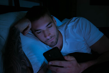 handsome man looking at his mobile phone in bed at night with glow of phone lighting his face