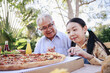 Asian retirement grandfather and pretty granddaughter enjoying to eating pizza together in home garden. Happy senior life after retirement with family concept.