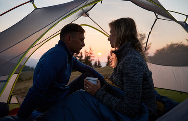 Wall Mural - Silhouette of family pair, who relaxing in tent early morning after cold mountain night on background of pink shadows sky during sunrise. Mountain recreation in camp.