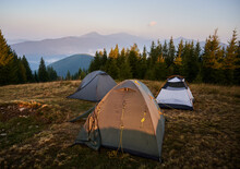 Tourist Camp With Three Tents Set Up On Meadow Near Forest. Morning Sunlight On Tent And Surrounding Nature. Silhouettes Of High Mountains On Background.