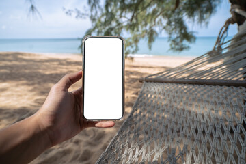 Wall Mural - Hand holding mock up mobile with white sceen while laying on hammock swing between trees on the beach.