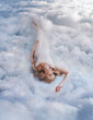 Young beautiful happy blonde woman lies in soft bed on white clouds in sky. Fantasy girl sleeping beauty. Goddess resting dreaming enjoying life at top of heaven Olympus. Divine light lady angel