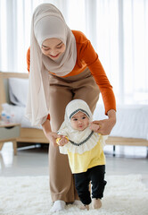 A beautiful Asian Muslim mother try to help the Muslim baby in a hijab dress for first walking on floor with love and care. Family and relationship concept