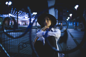 Poster - Young female in casual clothing and a hat posing near city lights at night