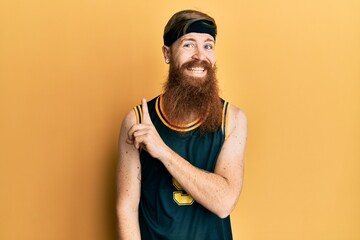 Sticker - Redhead man with long beard wearing basketball uniform with a big smile on face, pointing with hand and finger to the side looking at the camera.