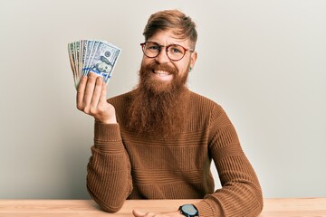 Sticker - Young irish redhead man holding dollars sitting on the table looking positive and happy standing and smiling with a confident smile showing teeth