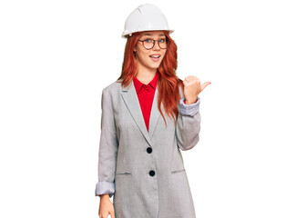 Wall Mural - Young redhead woman wearing architect hardhat smiling with happy face looking and pointing to the side with thumb up.