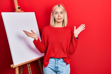 Poster - Beautiful blonde woman standing by painter easel stand clueless and confused with open arms, no idea and doubtful face.