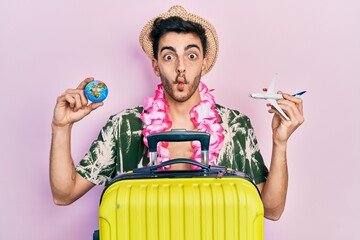 Wall Mural - Young hispanic man wearing summer style and hawaiian lei holding world ball and plane toy making fish face with mouth and squinting eyes, crazy and comical.