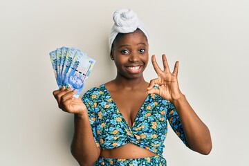 Wall Mural - Young african woman wearing hair turban holding south african 100 rand banknotes doing ok sign with fingers, smiling friendly gesturing excellent symbol