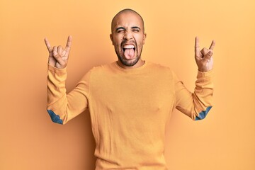 Wall Mural - Hispanic adult man doing rock gesture over yellow background sticking tongue out happy with funny expression.