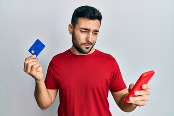 Young hispanic man holding smartphone and credit card skeptic and nervous, frowning upset because of problem. negative person.